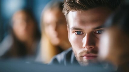 A close up of a serious young professional deeply engaged with his computer screen in a collaborative workspace. The blurred background highlights teamwork and a dynamic office atmosphere