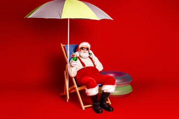 Santa Claus relaxing on a deck chair under a colorful umbrella with a drink and inflatables against a red backdrop for a festive holiday scene