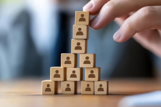 Building the Foundation: A hand carefully places a block atop a pyramid of wooden blocks, each etched with a simple figure, symbolizing the building of a strong team and human resource growth.
