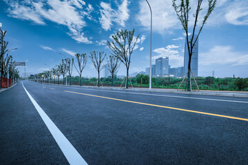 Naklejka premium Empty winding asphalt road through a modern urban park area with city buildings in the background under a blue sky.
