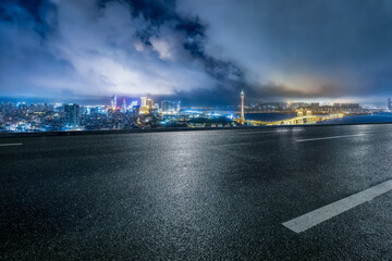 Empty asphalt road overlooking the illuminated modern city skyline and bridge at night.