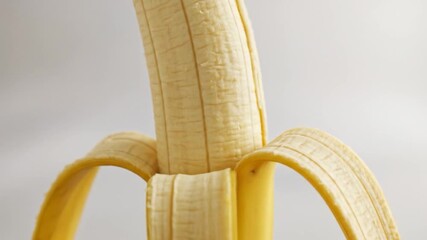 A peeled banana, revealing its creamy flesh, sits against a soft white background. The textured peel arcs outwards - Powered by Adobe