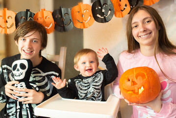 Fototapeta premium Brothers and sister in skeleton print pajamas celebrate Halloween. Children sit together, a teenage girl holds a jack-o'-lantern. Brother and sister, teenagers, younger brother, baby.