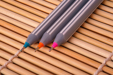 Close-up of three colorful ballpoint pens (blue, orange, pink) placed on a bamboo surface. Representing creativity, stationery, office supplies and writing tools in vibrant contrast.