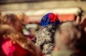 A young child wearing a blue hat sits on a parent’s shoulders, eagerly awaiting the arrival of...