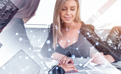 Two businesswomen collaborating at desk with digital network graphics overlay, in bright modern office setting, representing teamwork and digital strategy concept