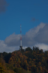 Fototapeta premium A tall red and white communications tower stands on a forested hilltop under a partly cloudy blue sky during the daytime.