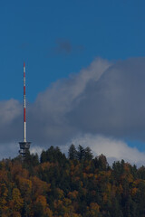 Fototapeta premium A tall red and white communications tower stands on a forested hilltop under a partly cloudy blue sky during the daytime.