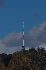 Fototapeta premium A tall red and white communications tower stands on a forested hilltop under a partly cloudy blue sky during the daytime.