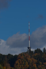 Obraz premium A tall red and white communications tower stands on a forested hilltop under a partly cloudy blue sky during the daytime.