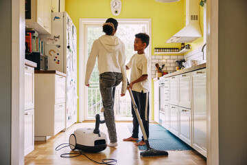 Rear view of girl walking near brother holding vacuum cleaner in kitchen at home