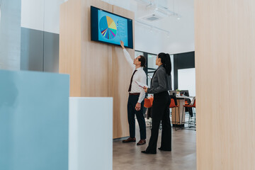 Fototapeta premium Two colleagues review a colorful dashboard on a wall screen inside a bright, modern office. They discuss data, pie charts, and strategy, highlighting teamwork, diversity, and business collaboration.