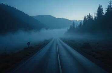 Empty asphalt road disappears into thick blue fog. Dark mountains covered with pine forest flank highway. Serene twilight landscape creates mysterious travel adventure scene. Peaceful nature view