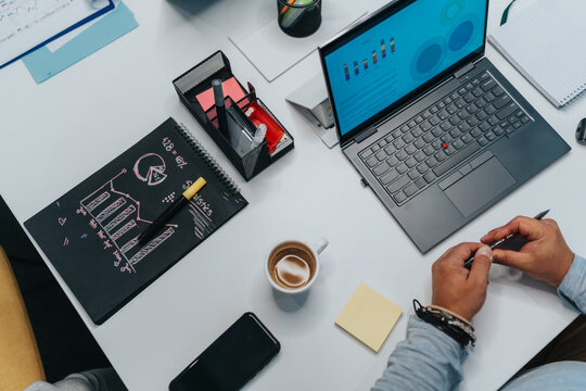A bustling desk scene featuring a laptop with charts, a chalkboard notebook, coffee, and hands at work. Ideal for topics on teamwork, planning, and modern office collaboration.