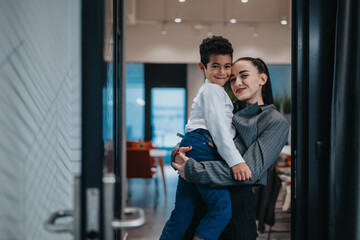 A mother holds her smiling son in the doorway of a contemporary office space, capturing warmth, affection, and family bond in a professional environment.