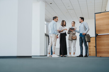 A diverse group of colleagues stands in a bright office corridor, sharing a tablet and notes as...