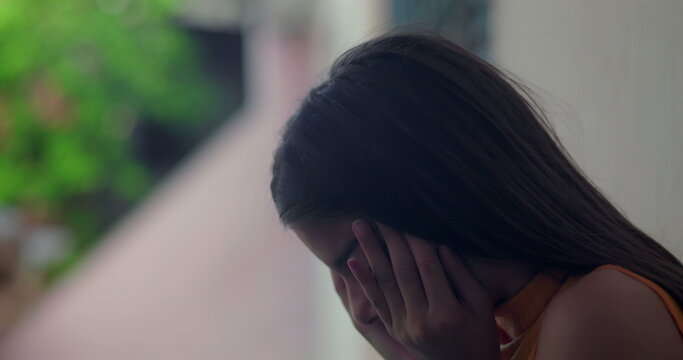Young girl with brown hair, covering her face with hands, eyes closed, reflecting deeply while leaning against a wall outdoors, emotional moment in a peaceful urban setting
