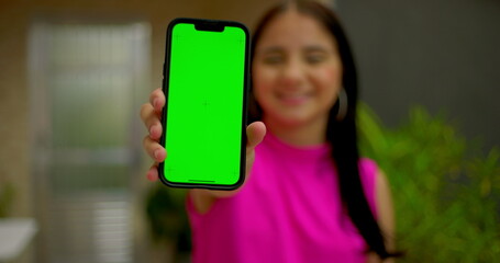 Young woman holding smartphone with green screen chroma key close to the camera, smiling confidently, showcasing blank screen for customizable content in an outdoor home setting