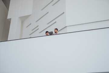Two professionals stand at a white balcony, sharing ideas over a laptop in a sleek, modern...