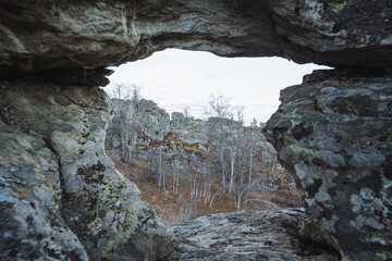 Natural rock window revealing forest basin below and distant ridge, textured arch with lichen framing panoramic vista, soft cool light and layered composition evoke calm remote landscape