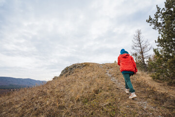 Fototapeta premium Young boy climbs rugged slope, Young child wearing colorful clothing moves across rugged terrain, Children in vibrant attire ascend challenging hill under overcast sky with determination
