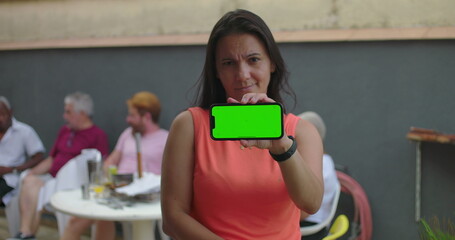 Woman holding smartphone with green screen directly towards camera, chroma key, neutral expression, standing outdoors with casual group conversation in blurred background
