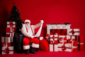 Santa reads a long list among gifts beside a Christmas tree against a bold red backdrop with...