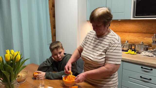 With a pile of fresh oranges on the table, a grandmother guides her grandson's hands as they press the fruit onto a manual juicer, creating healthy homemade juice.