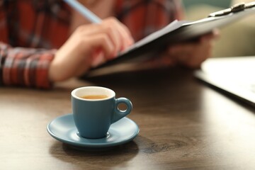 Coffee break. Cup of aromatic drink on wooden table and woman with clipboard indoors, selective focus