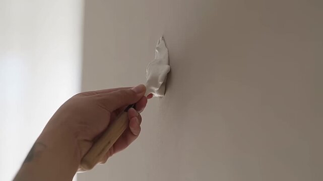 Close up of a hand applying filler to a small wall hole using a spatula in soft daylight. Smooth, even motion with a minimalist composition and neutral wall tone, captured in 4K detail.