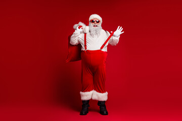 Santa in red suit with bag and suspenders stands against red background ready for holiday fun and...