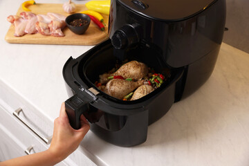 Woman cooking chicken drumsticks and vegetables in air fryer at white marble table, closeup