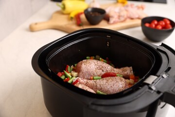 Air fryer basket with raw chicken drumsticks, vegetables and spices on white marble table, closeup