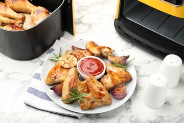 Delicious grilled chicken wings, sauce and spices near air fryer with cooked drumsticks on white marble table, closeup