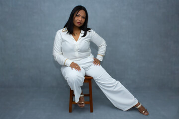Beautiful brown skin woman in white clothes, sitting on a stool with one straight leg and hand on...