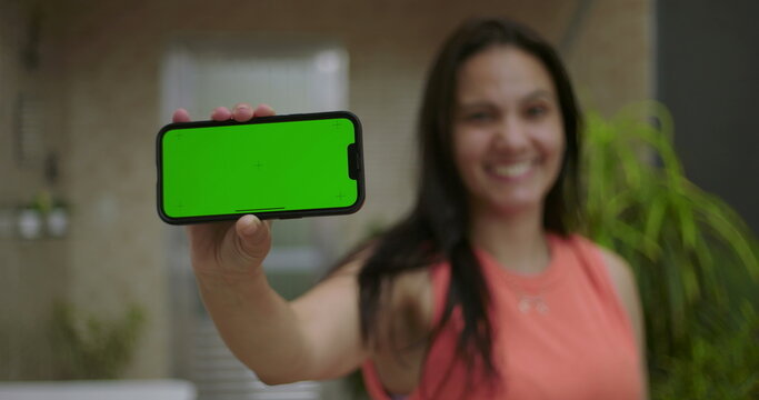 Woman holding smartphone with green screen and chroma key close to the camera, blurred background with outdoor setting, smiling and presenting device for customizable content - Powered by Adobe