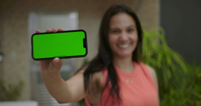 Woman holding smartphone with green screen and chroma key close to the camera, blurred background with outdoor setting, smiling and presenting device for customizable content