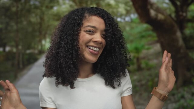 Young woman covers face with both hands on sunlit forest path lined with tall trees and green foliage; shyness.