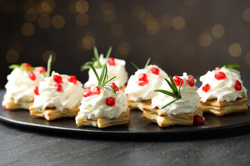 Sweet canapes with puff pastry, whipped cream, pomegranate seeds and rosemary on dark textured table against blurred lights, closeup. Christmas food