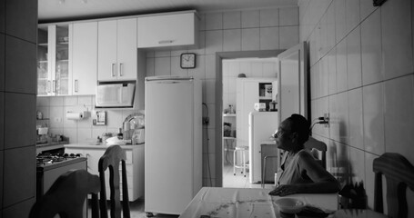 Elderly woman sitting at a dining table in a tiled kitchen, gazing thoughtfully towards the light,...