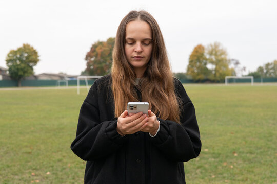 A woman with long hair is focused on her phone while standing on a soccer field surrounded by trees. The sky is overcast and the grass is lush