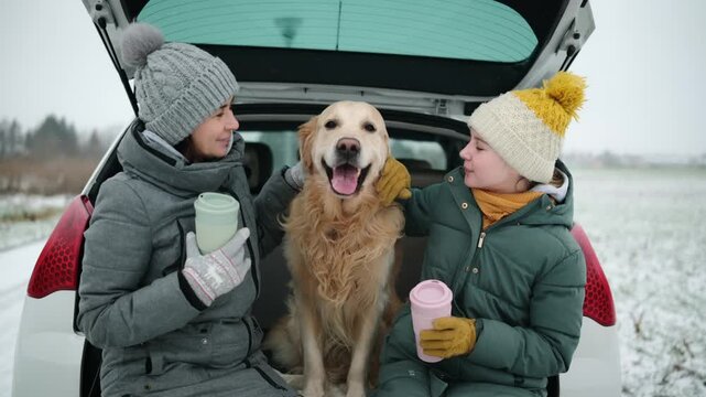 Mother And Daughter Happy With Dog In Car Trunk At Winter Road Rest Stop Drinking Hot Tea