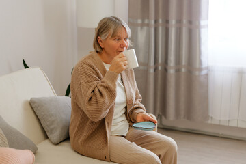 A woman sits comfortably on a sofa, sipping a warm drink and holding a coaster, enjoying a peaceful moment in her cozy living room with soft lighting