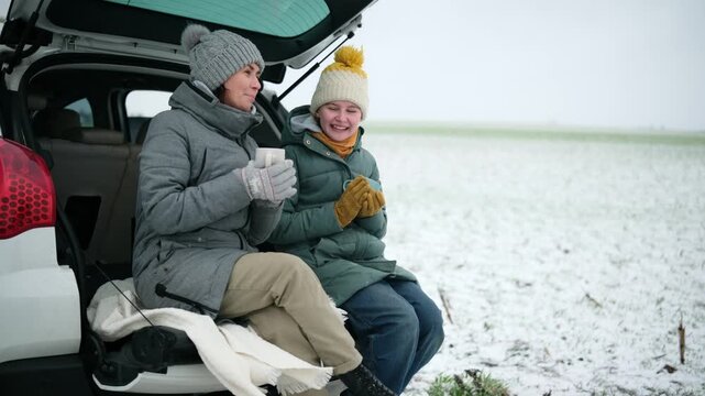 Happy Mother And Daughter Drinking Tea While Sitting In The Trunk Of A Car On A Rural Road During A Winter Travel Stop