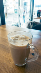 Vertical shot of frothy coffee in glass mug on wooden table inside cozy cafe, perfect for lifestyle, hospitality, and food photography
