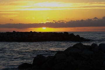 Sunset over calm ocean waves, with rocky shoreline silhouetted against vibrant orange and yellow sky, creating a serene and tranquil evening atmosphere by the water