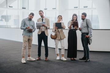 Group of colleagues and friends stands in a bright, modern corridor, examining tablets and papers....