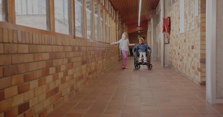 Two classmates rolling wheelchair and walking with backpack in school hall, with fire hose cabinet