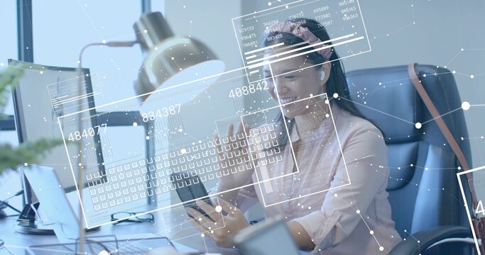 Holding smartphone while analyzing data overlays, woman wearing pink blouse, earbuds at office desk