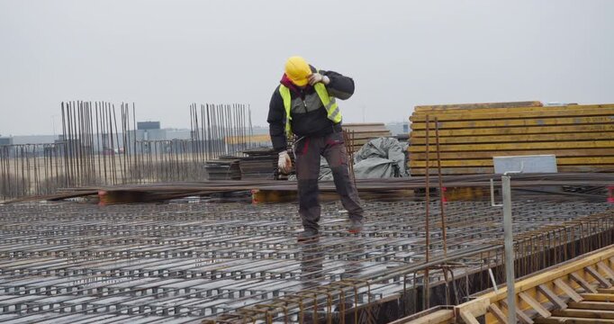 Construction worker in high-visibility vest and helmet adjusts steel reinforcement bars on a concrete slab, preparing the structure for pouring. Precision and safety essential for building integrity.